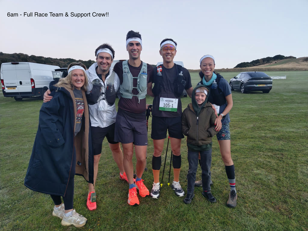 Six people standing arm in arm on grass by the support vans before the race, wearing white headbands, one with race number 105-1 pinned on.